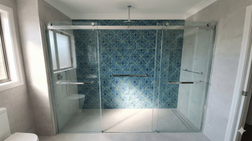 A spacious alcove shower installed across the full width of a bathroom wall, designed to replace a standard bathtub. The layout features sliding glass doors to save space and blue geometric feature tiles on the back wall, demonstrating a modern bath-to-shower conversion.