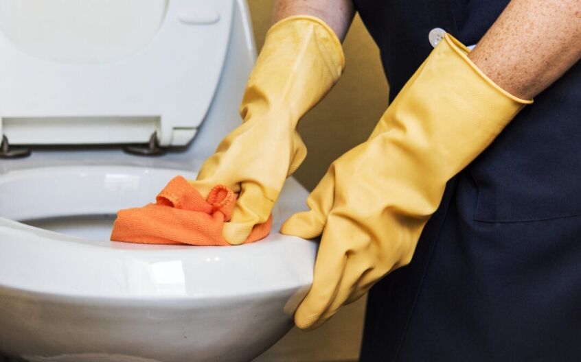 Person wearing gloves cleaning a raised toilet seat with disinfectant spray and cloth.