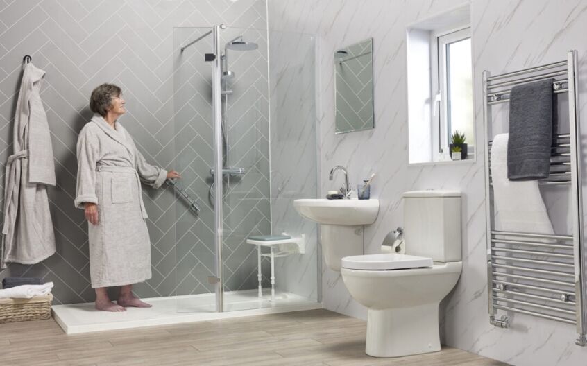 Older woman in bathrobe standing beside level-access walk-in shower with fold-down seat, grab rail, and modern fittings in accessible bathroom