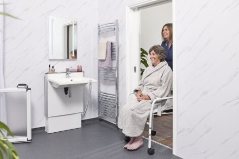 Older woman in a wheelchair in a spacious accessible bathroom, helped by a carer