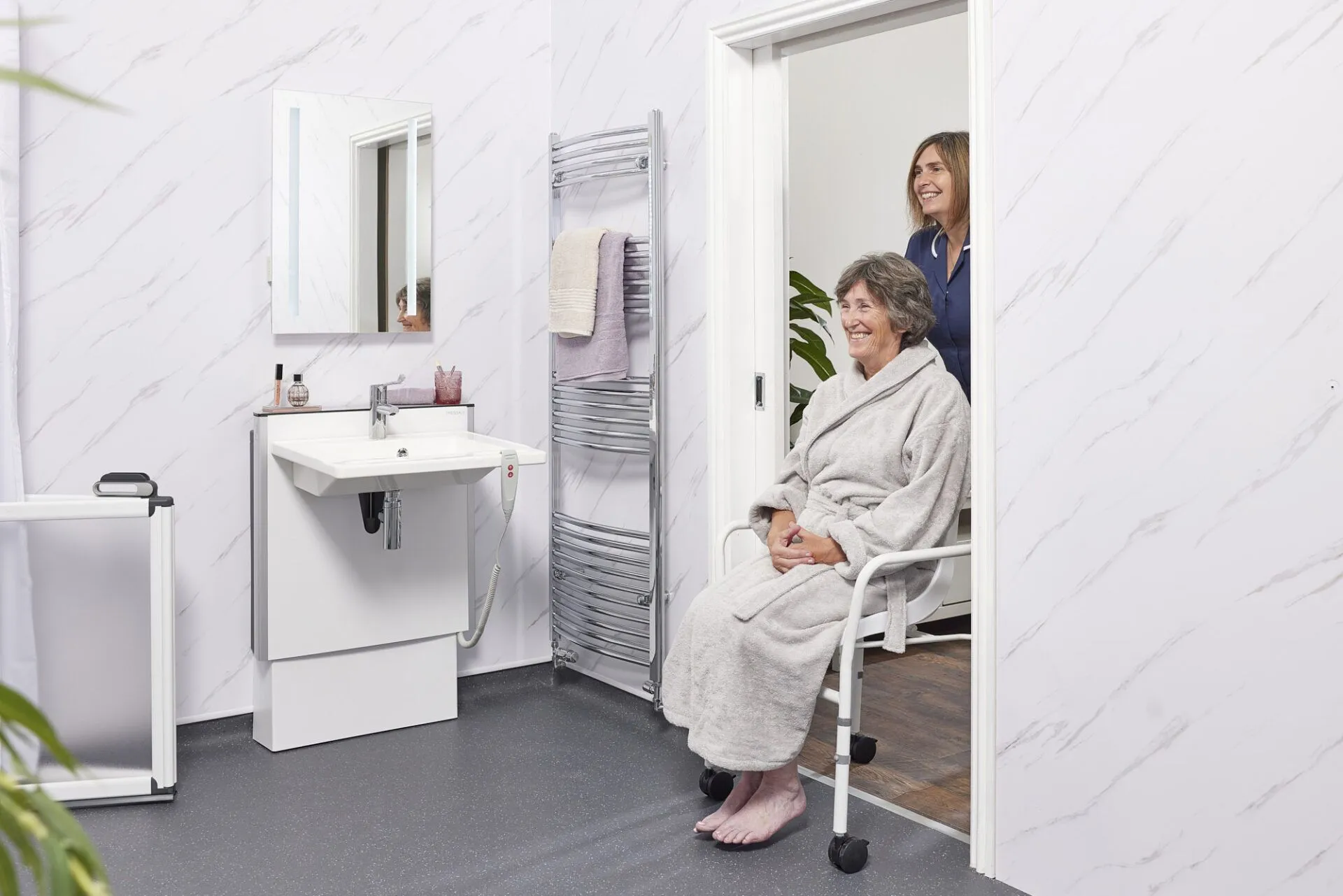 Elderly woman in a mobility shower chair smiling as a carer assists her in an accessible bathroom with a height-adjustable sink and grab rails.