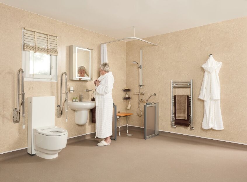 Accessible wet room with beige walls, raised toilet and grab rails, with a woman standing at the basin