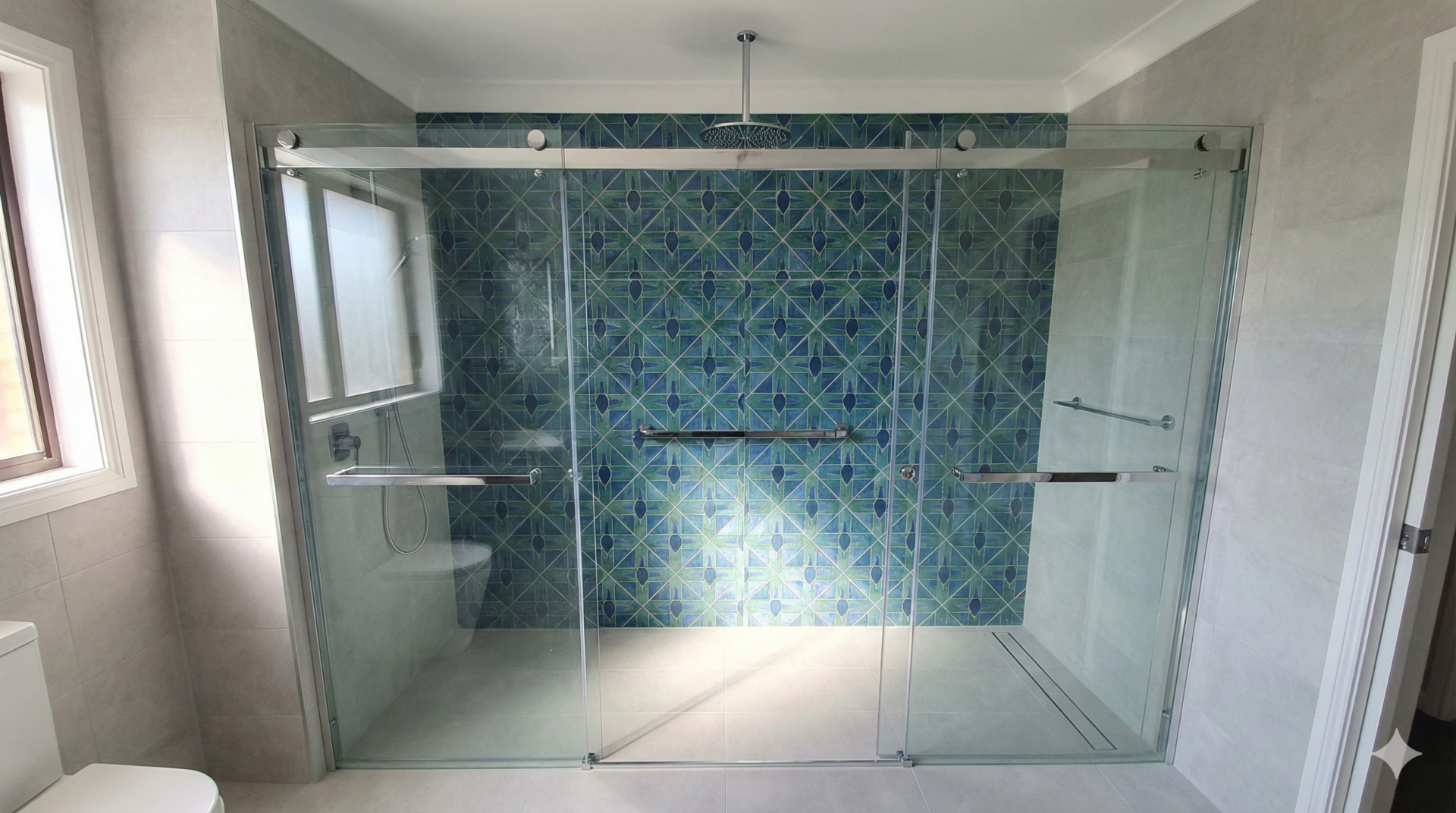 A spacious alcove shower installed across the full width of a bathroom wall, designed to replace a standard bathtub. The layout features sliding glass doors to save space and blue geometric feature tiles on the back wall, demonstrating a modern bath-to-shower conversion.
